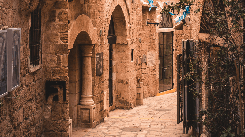 Historic stone alleyway with archways and a cozy walkway in an old city, adorned with blue and white banners.